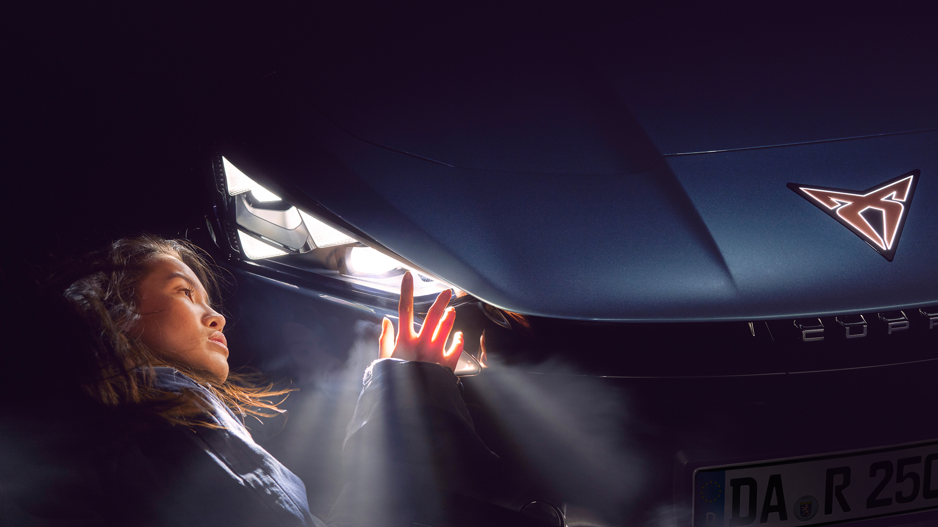 Frau in Nahaufnahme blickt auf den eingeschalteten Frontscheinwerfer eines CUPRA Tavascan, im Vordergrund das beleuchtete CUPRA Logo auf der Motorhaube